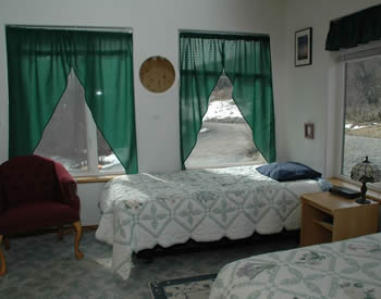 Shows room with windows on two walls and two beds with green and white bedspreads and a burgundy chair.